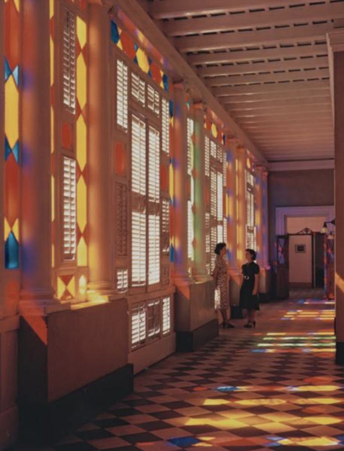 This image depicts deeply saturated stained glass windows leaking into the light in a building in Puerto Rico. Two women stand in the background.