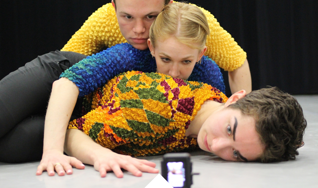 Three dancers lie atop each other onstage staring intensely at a strobe light